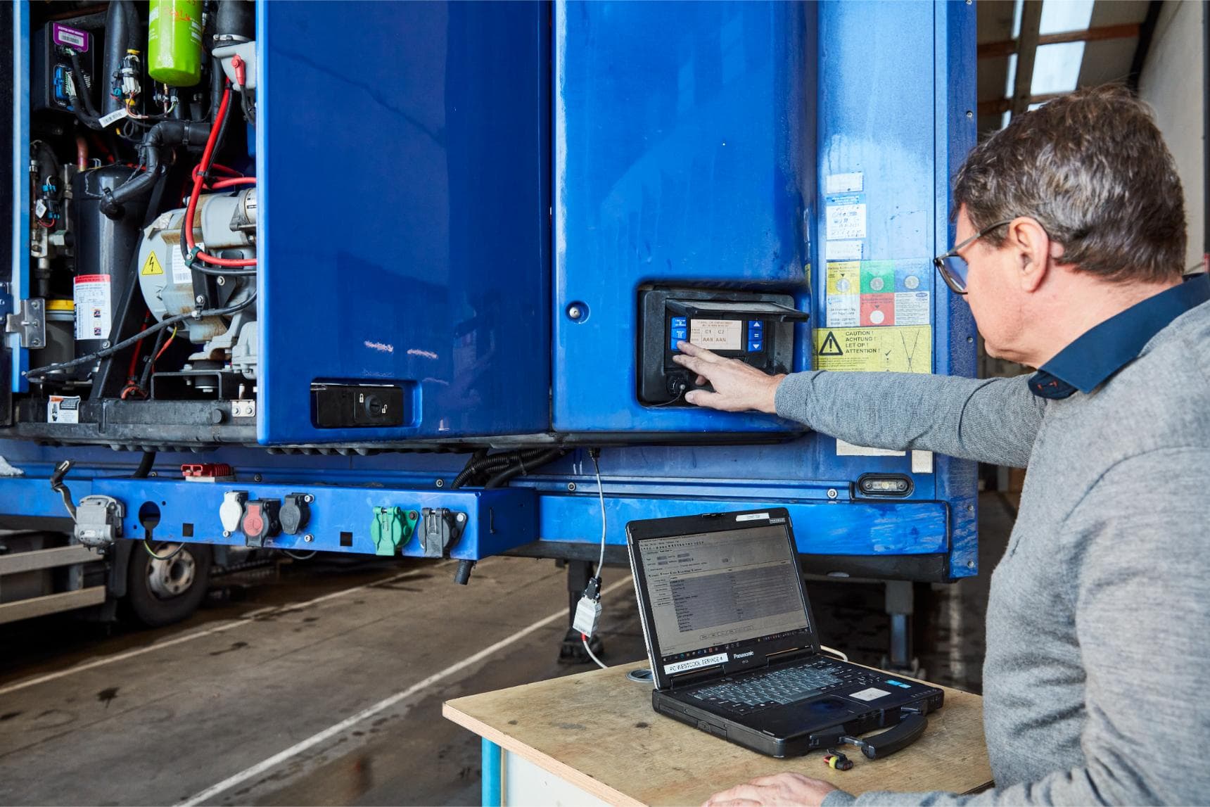 A technician reading out the control box of a refrigeration unit on a blue trailer with a laptop